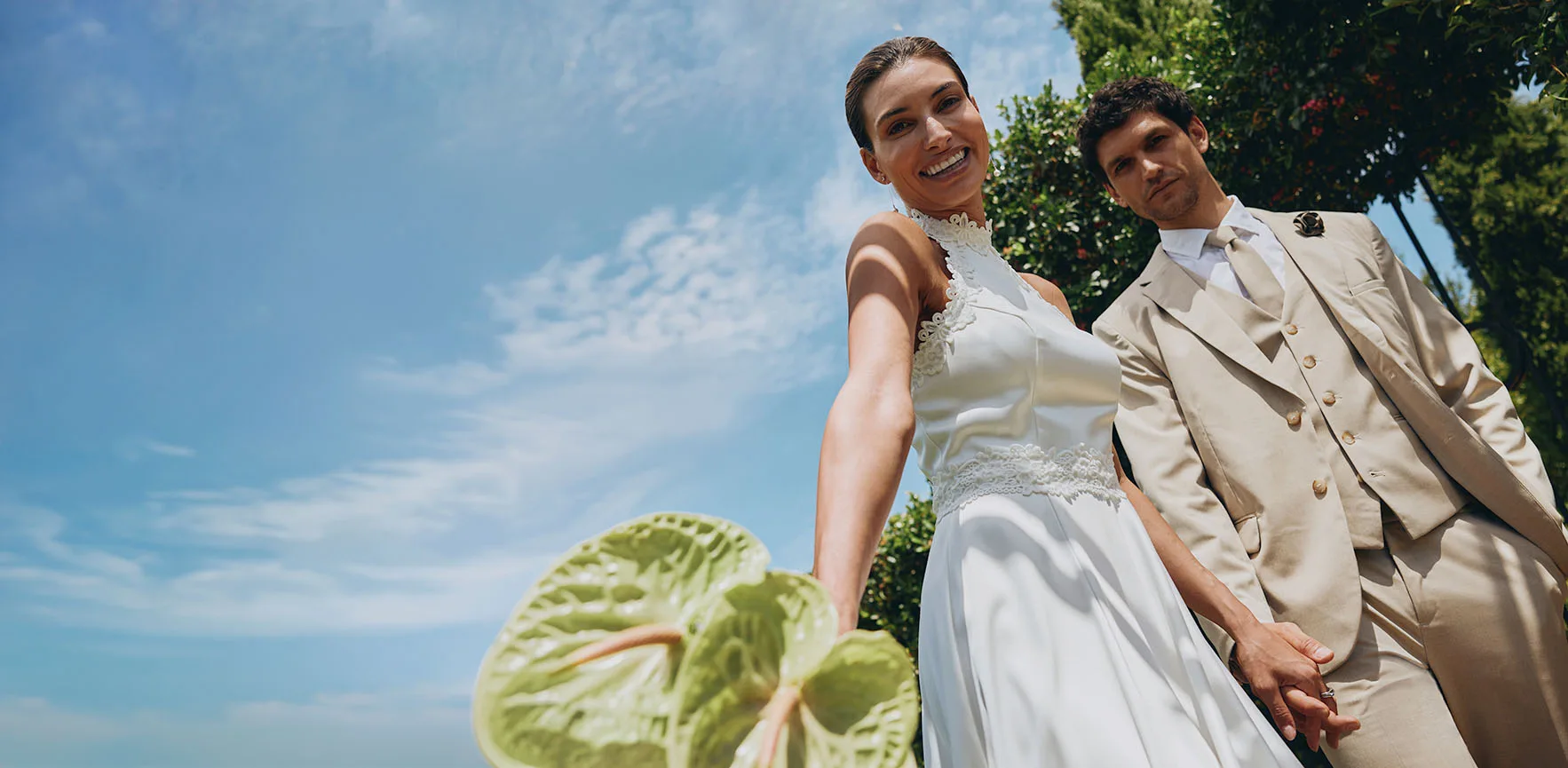 Elegáns menyasszony és vőlegény napsütésben: a menyasszony fehér ruhában, a vőlegény bézs színű öltönyben, napszemüveggel.